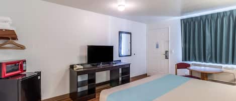 Habitación de hotel con cama, escritorio con televisor, microondas, refrigerador pequeño, espejo, ventana con cortinas y puerta.