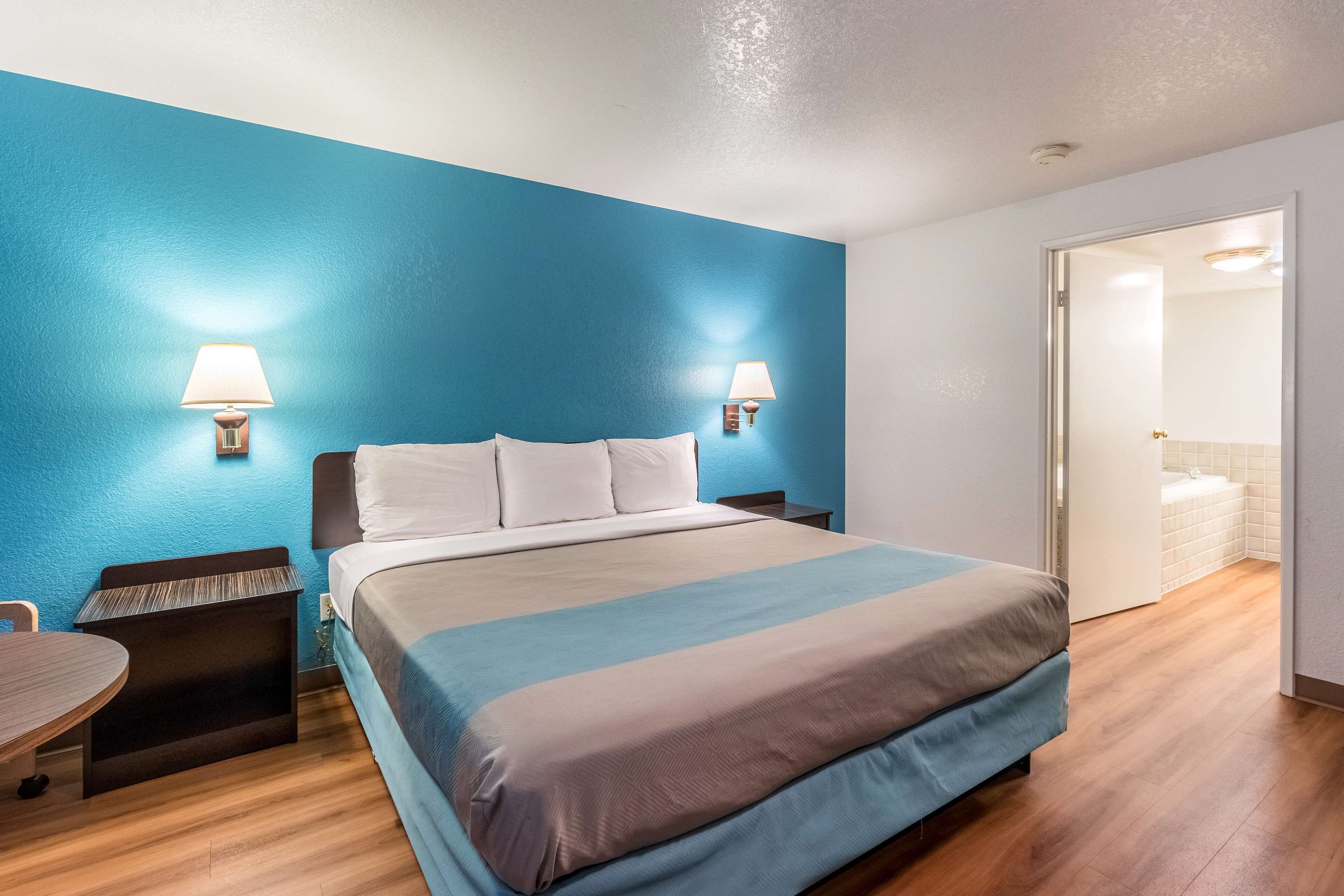 A hotel room with a large bed, two bedside lamps, a small table, and a door leading to a bathroom.