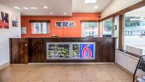 A hotel reception area with a wooden reception desk, framed pictures on the wall, and a large window providing a view of the outside. - Motel 6 Astoria, OR (Astoria)