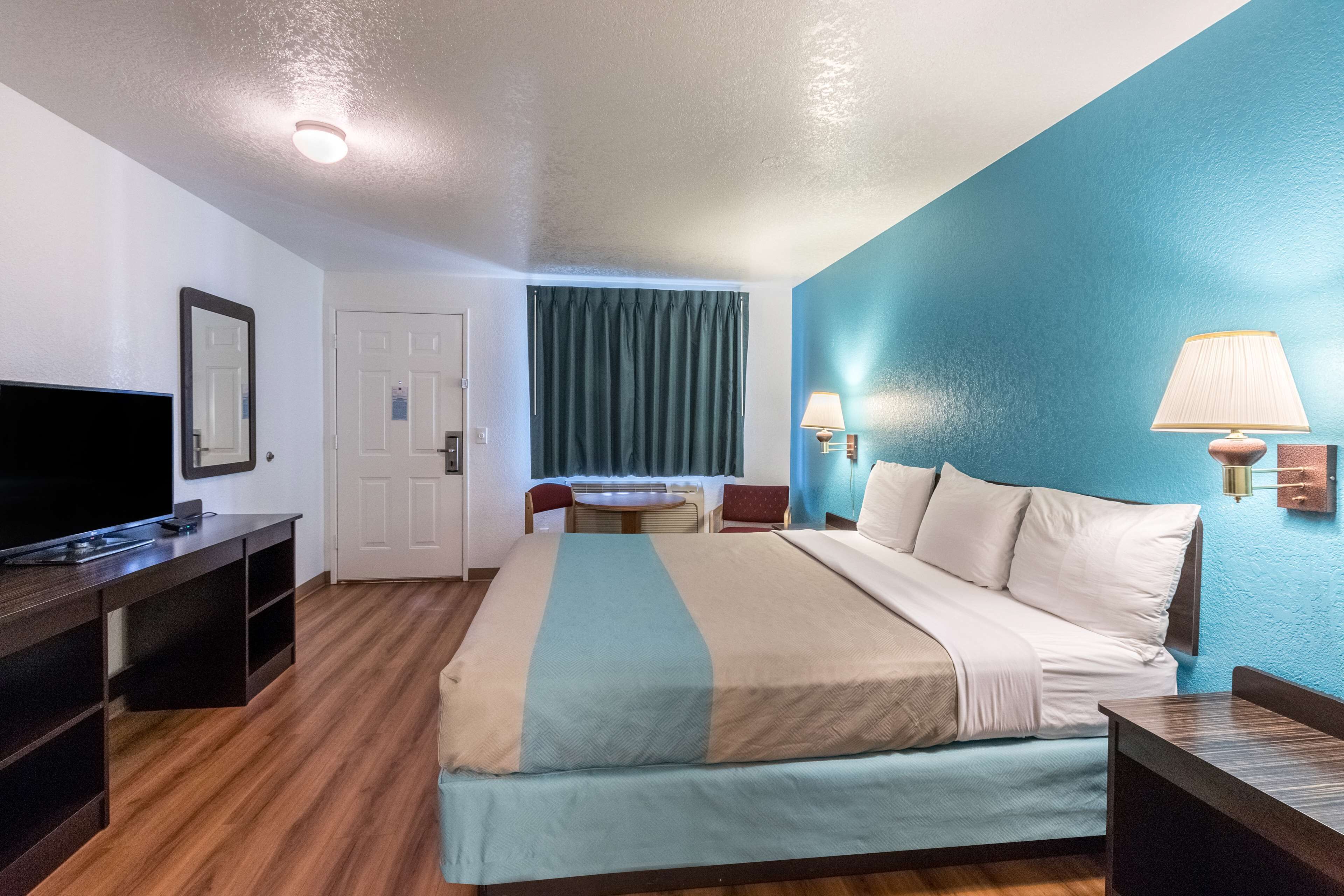 A hotel room with a bed, a desk with a TV, a table with chairs, a lamp, and a window with curtains.