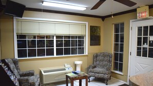 A room with two patterned armchairs, a small table with books, a window with blinds, a white door, and an air conditioning unit. - Claremore Motor Inn (Claremore)