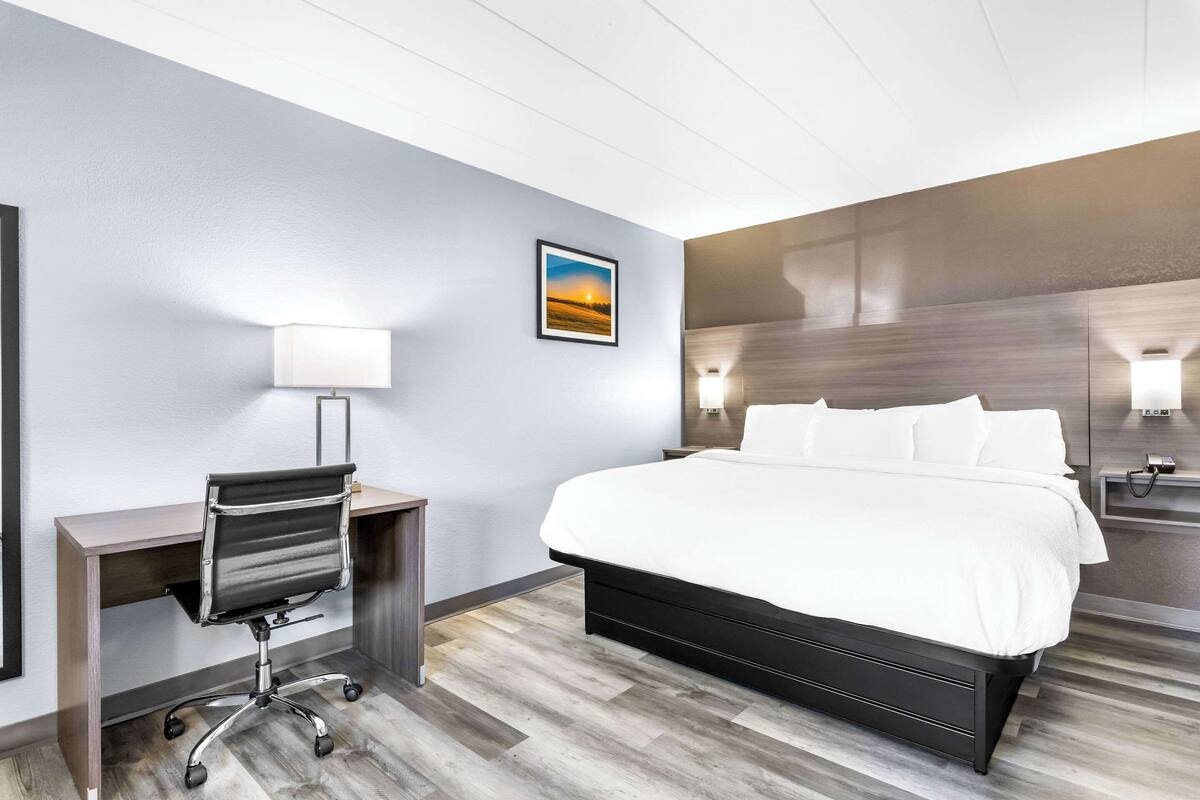 A hotel room with a bed, desk, chair, and a painting on the wall.