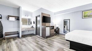 A hotel room with a bed, a desk, a chair, a TV, a mirror, a sink, and a shower.