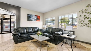 A modern living room with a black leather sofa, a coffee table, and a lamp. There are large windows providing natural light and a view of the outside.