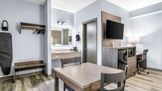 A modern hotel room with a wooden dining table, a small bench, a desk with a chair, a TV mounted on the wall, and a mirror above the sink.
