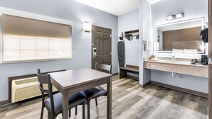 A hotel room with a dining table, chairs, a kitchenette, a large mirror, and a window with blinds.