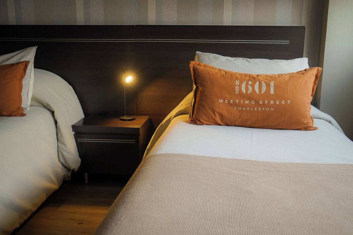 Habitación de hotel con dos camas, una mesita de noche con lámpara y una almohada con el texto '601 Meeting Street Charleston'.