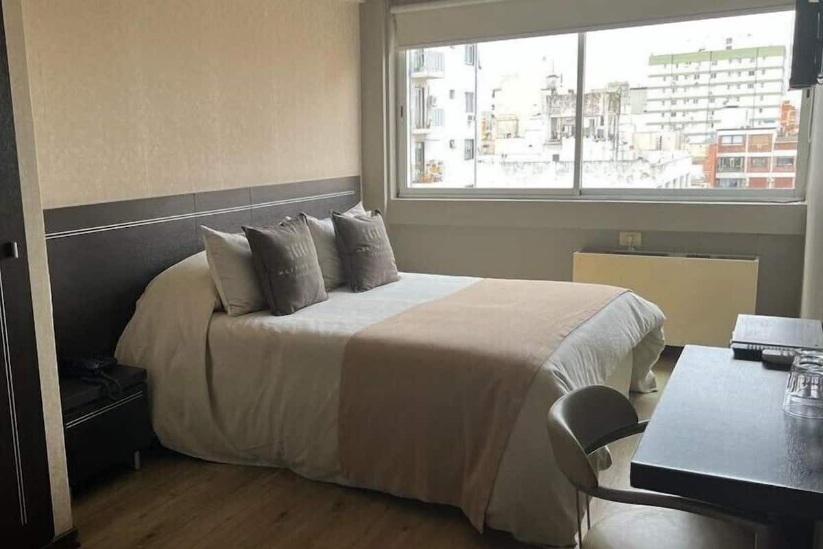 Habitación de hotel con cama, mesita de noche, silla y ventana con vista a edificios.