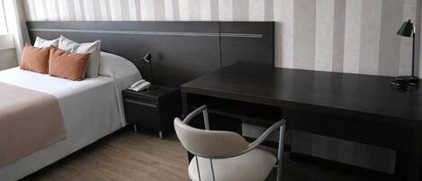 Una habitación de hotel con cama, mesita de noche, escritorio, silla y lámpara.