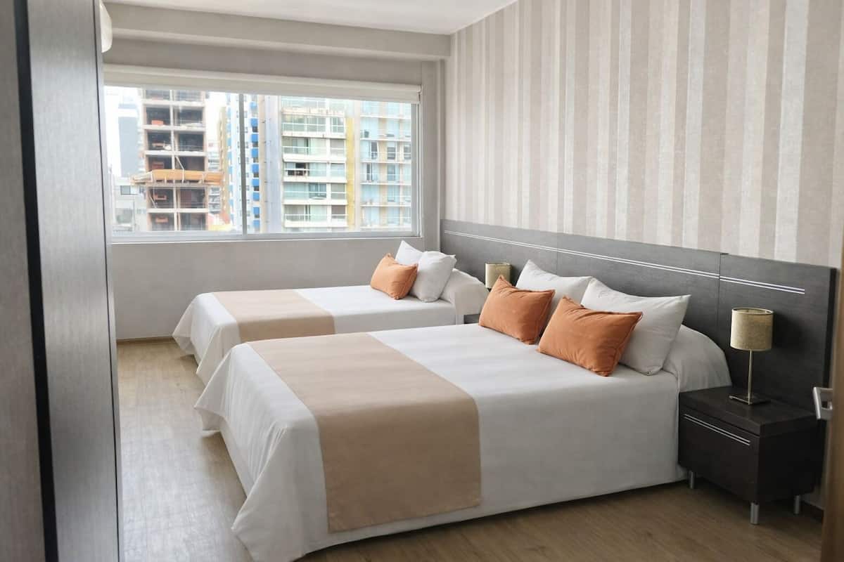Habitación de hotel con dos camas, una mesita de noche, una lámpara y ventana con vista a la ciudad.