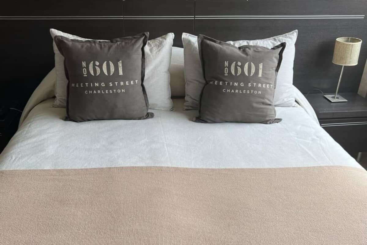 Una cama bien hecha con dos almohadas que dicen "N001 Meeting Street Charleston" y una manta beige.
