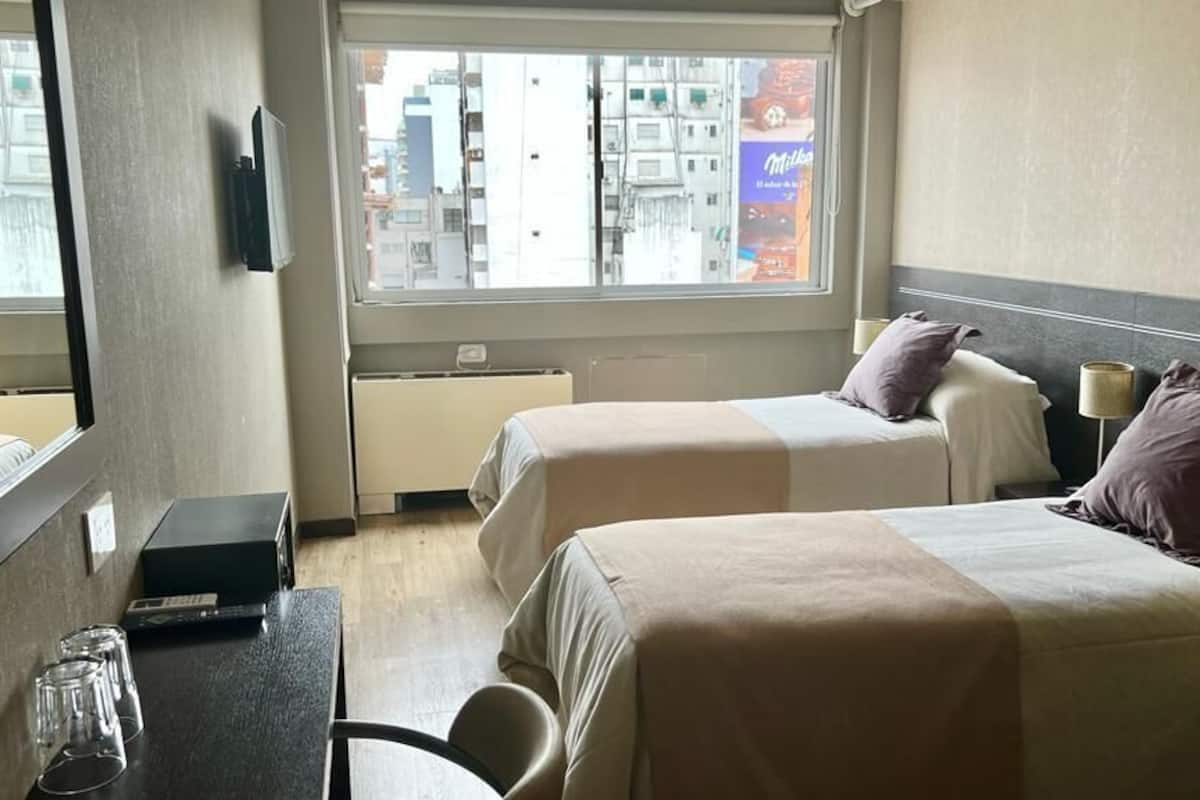 Una habitación de hotel con dos camas, un escritorio con computadora, un espejo y una ventana con vista a la ciudad.