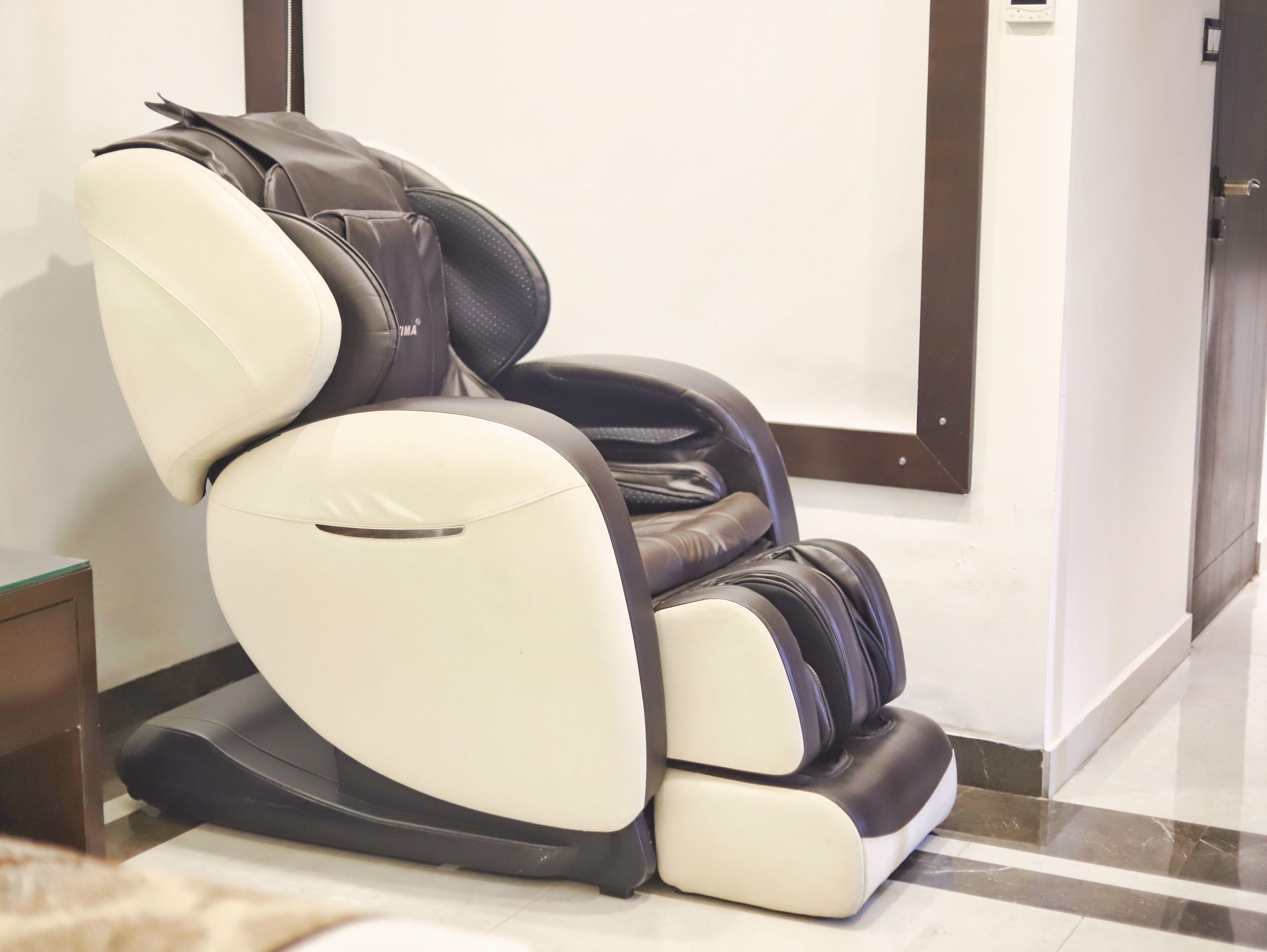 A massage chair with a reclining backrest and footrest, positioned in a room with a tiled floor and a door in the background.