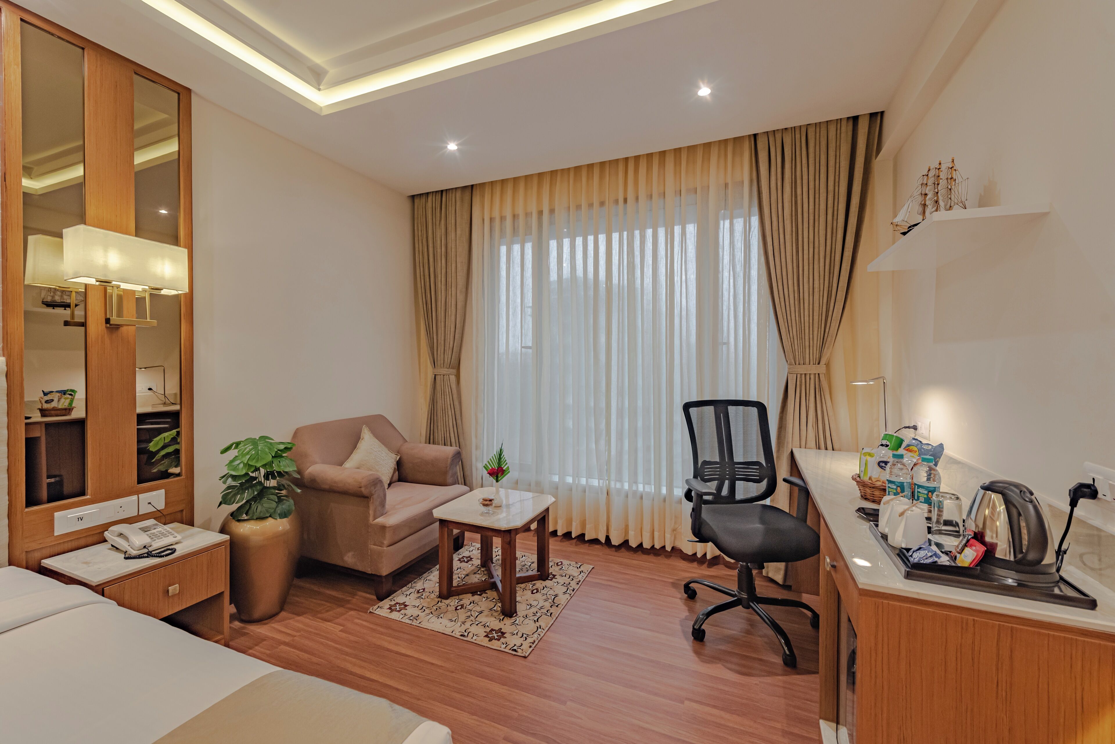A well-lit hotel room with a bed, a sitting area with a sofa and armchair, a small table with a lamp, a desk with a kettle and snacks, and a large window with curtains.