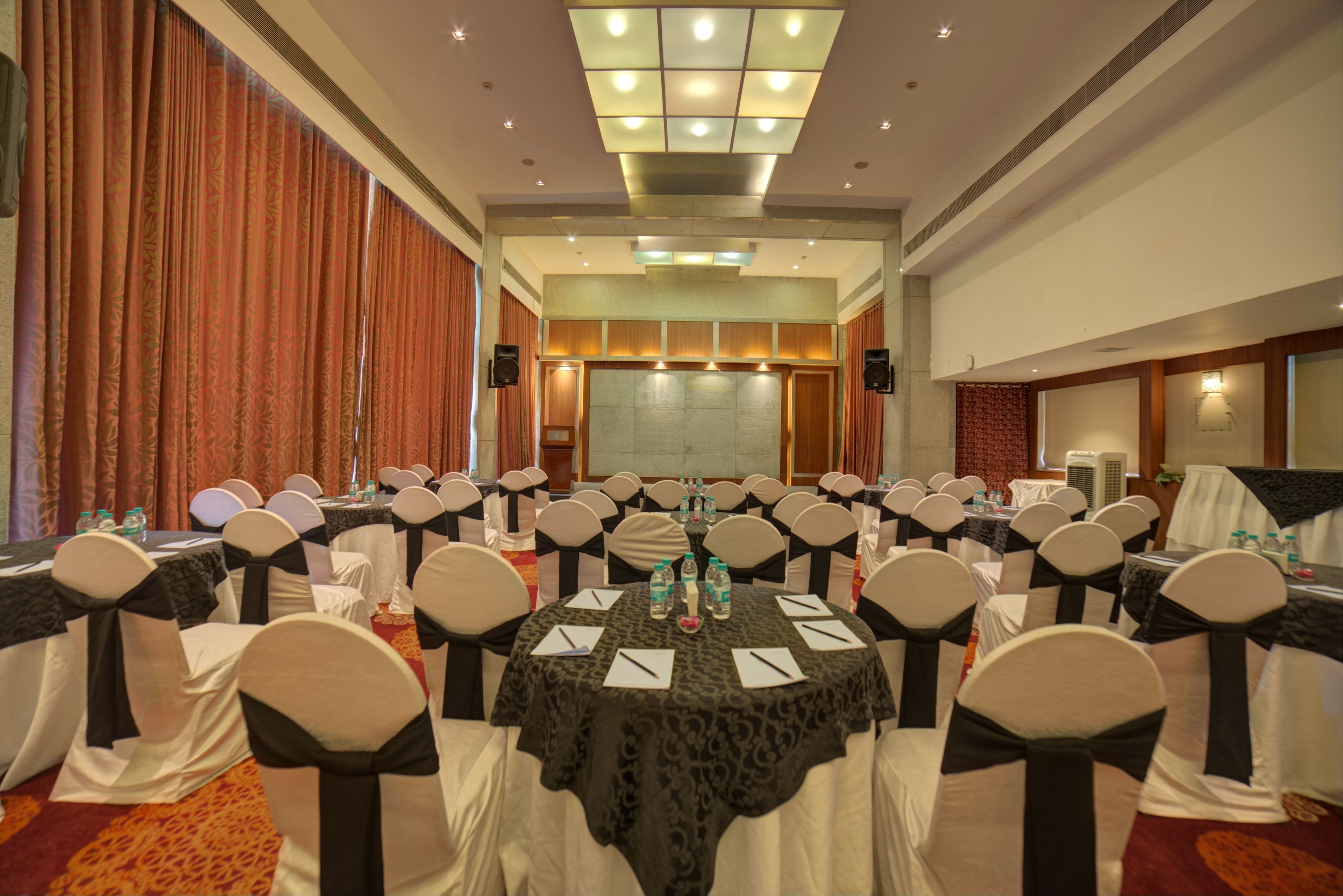 Une salle de conférence avec des tables rondes, des chaises blanches ornées de rubans noirs et un grand écran à l’avant.