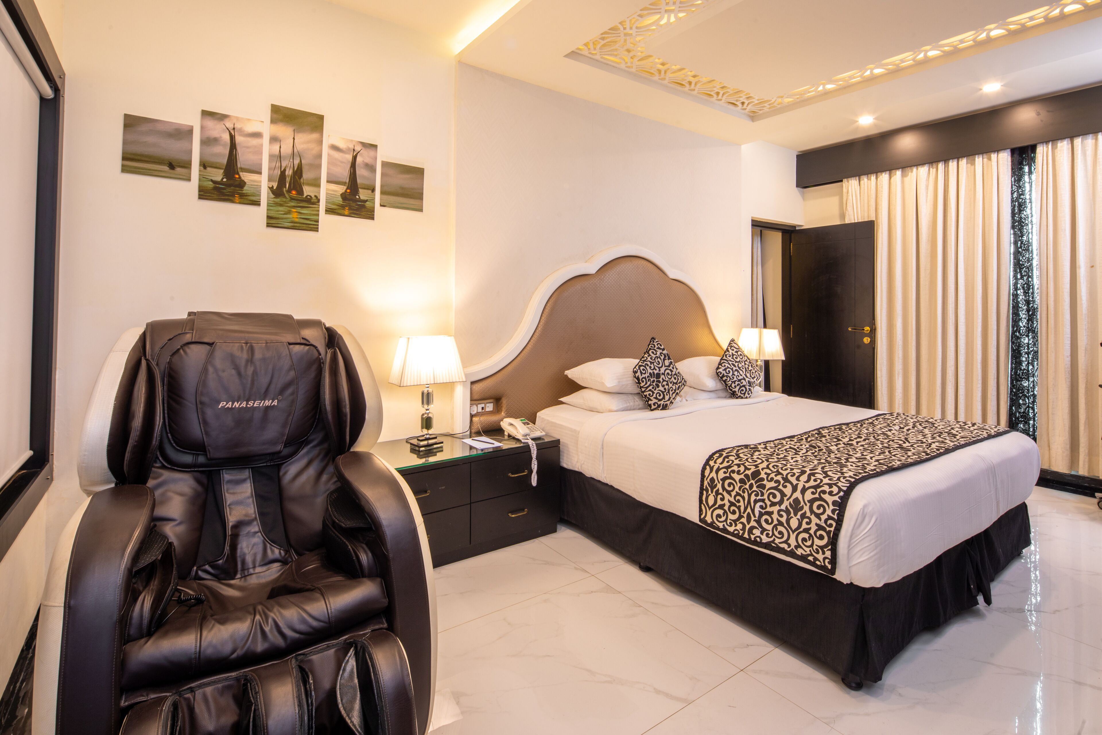 Une chambre d’hôtel équipée d’un fauteuil massant, d’un lit avec une literie à motifs, d’une table de chevet avec une lampe et d’œuvres d’art accrochées au mur.
