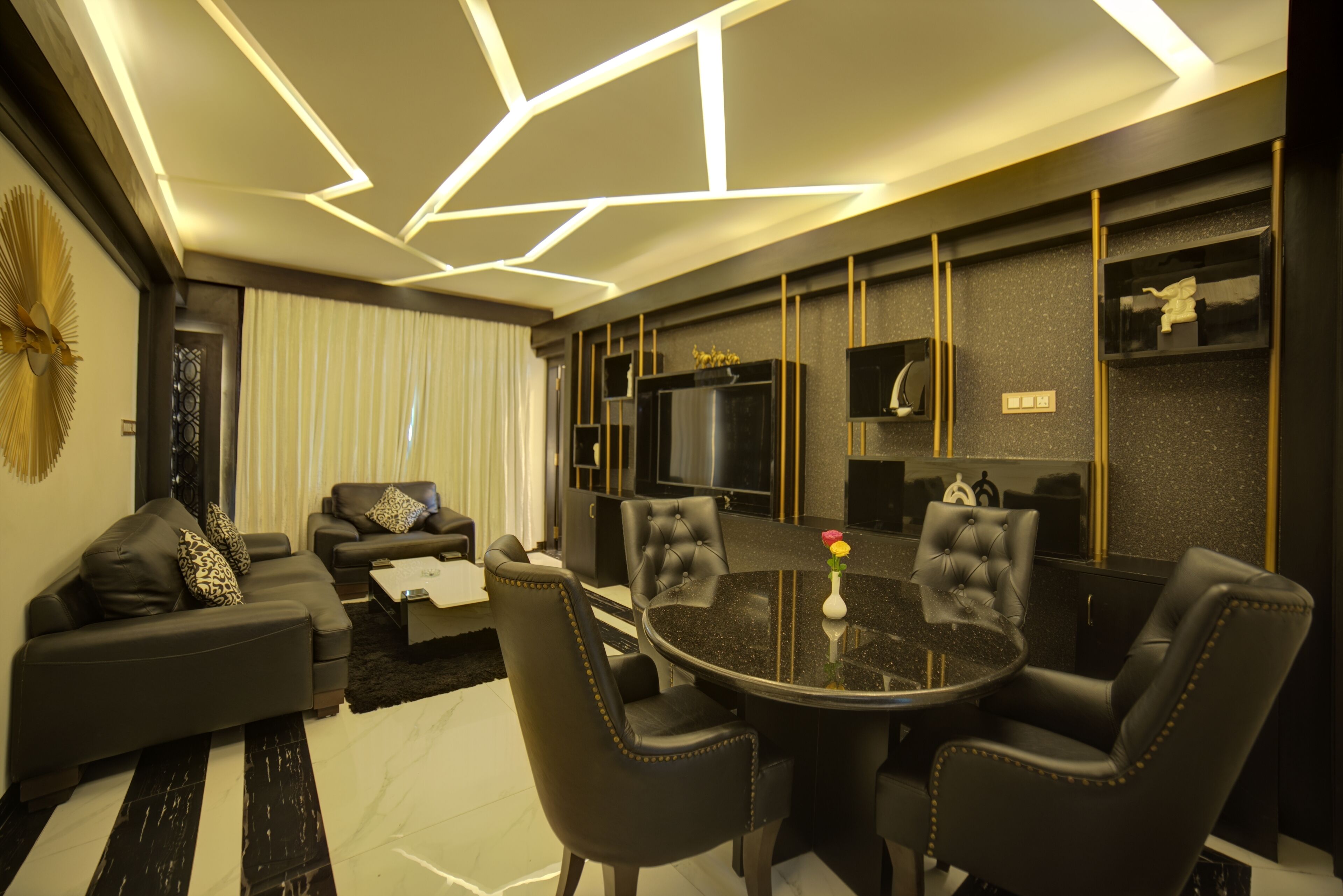 A modern living room with a black leather sofa, a round dining table with four chairs, and a flat-screen TV mounted on the wall.