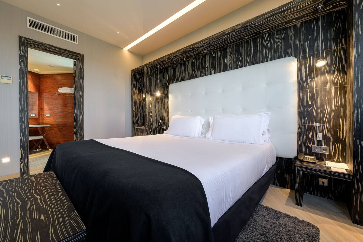 A modern hotel room with a large bed, a wooden accent wall, and a small bedside table with a lamp.