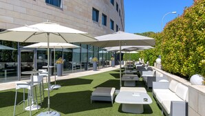 An outdoor patio with white furniture, including sofas and tables, under large umbrellas, and a building with large windows in the background.