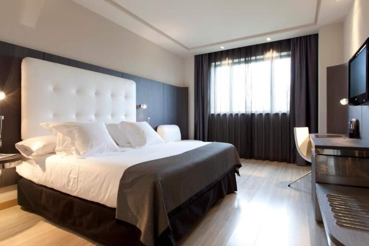 A modern hotel room with a large bed, a desk, a flat-screen TV, and a large window with curtains.