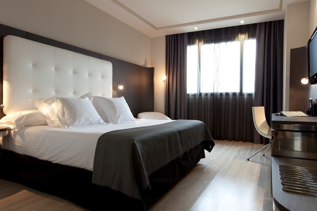 A hotel room with a large bed, a desk, a chair, a TV, and a window with curtains.