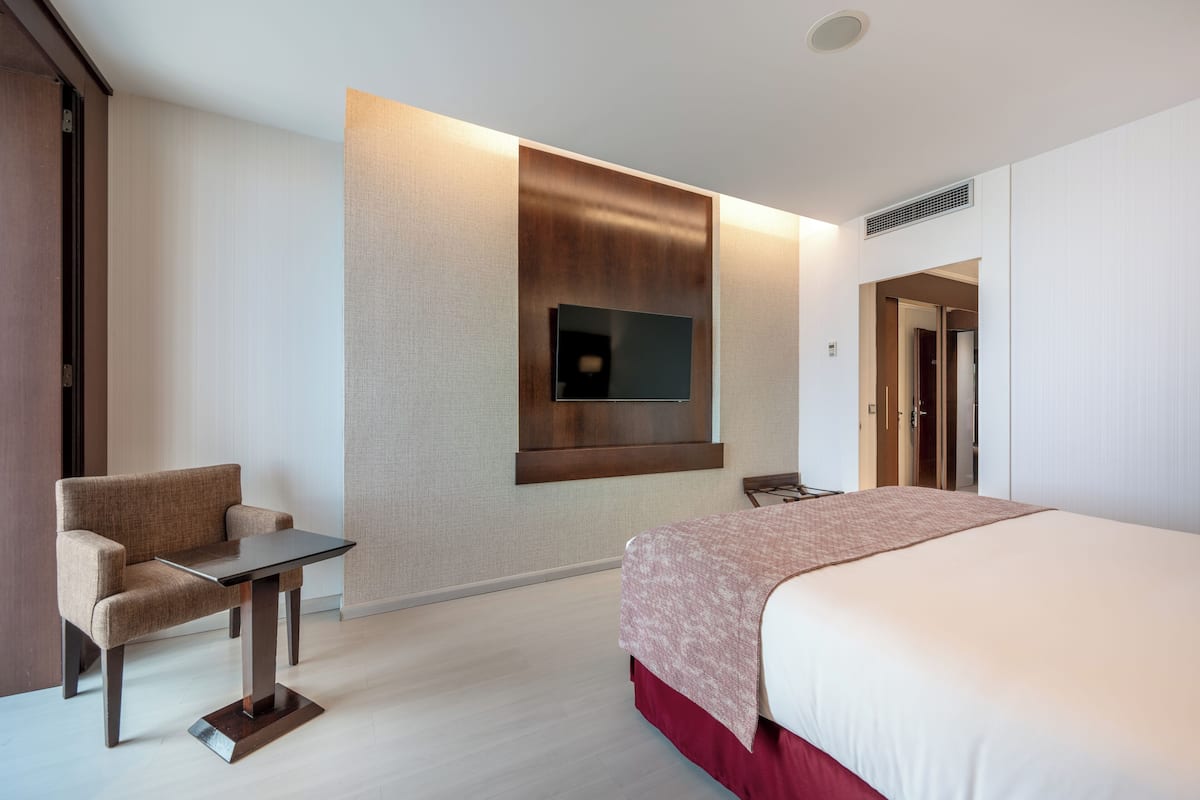 A hotel room with a bed, a desk, a chair, a TV mounted on the wall, and a door leading to another room.