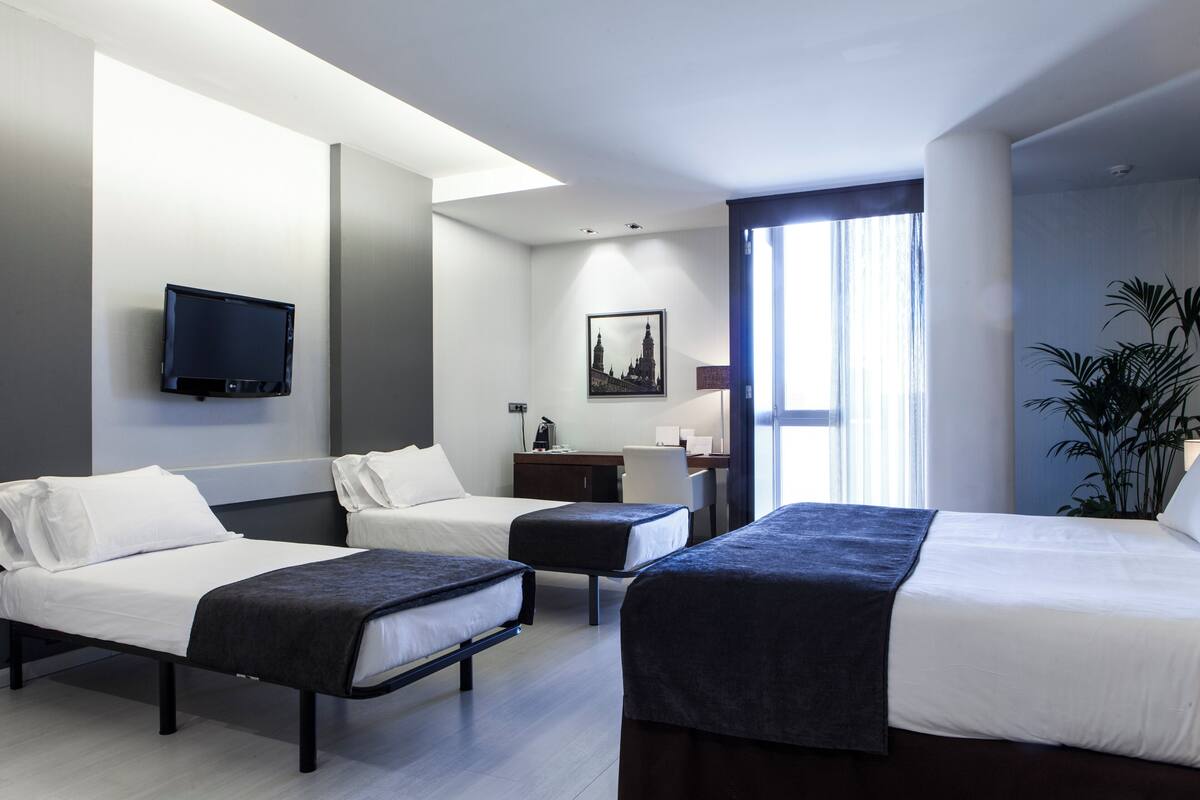 A hotel room with two single beds, a desk, a chair, a flat-screen TV mounted on the wall, and a large window with curtains.