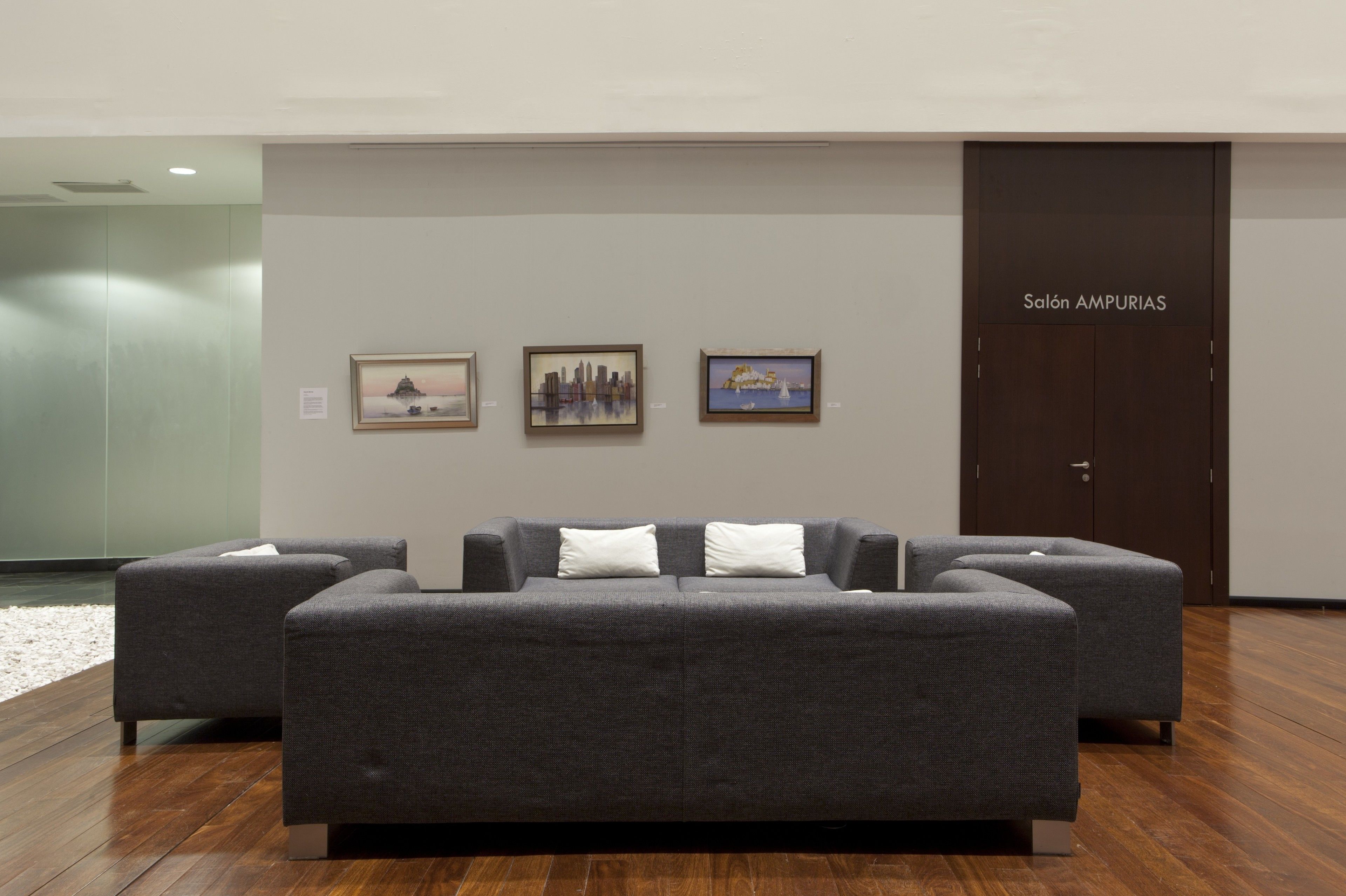 Um lobby moderno com um sofá cinzento, três obras de arte emolduradas na parede e uma porta com a inscrição "Salón AMPURIAS".