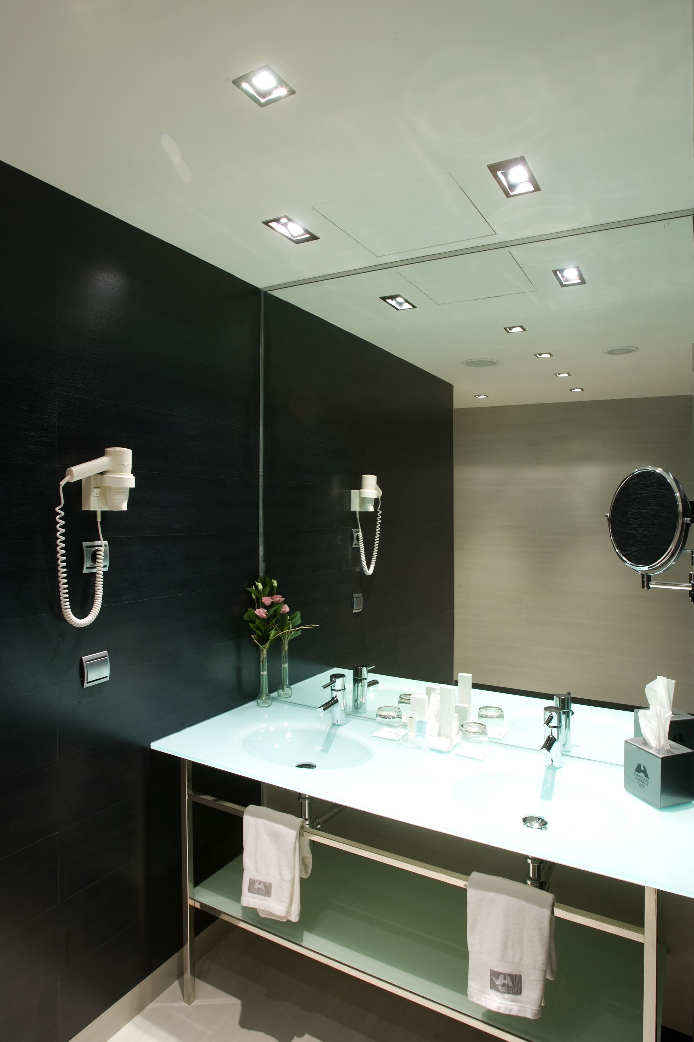 Une salle de bain moderne avec un grand miroir, deux sèche-cheveux fixés au mur, un lavabo blanc et une étagère en verre où sont posées des serviettes.
