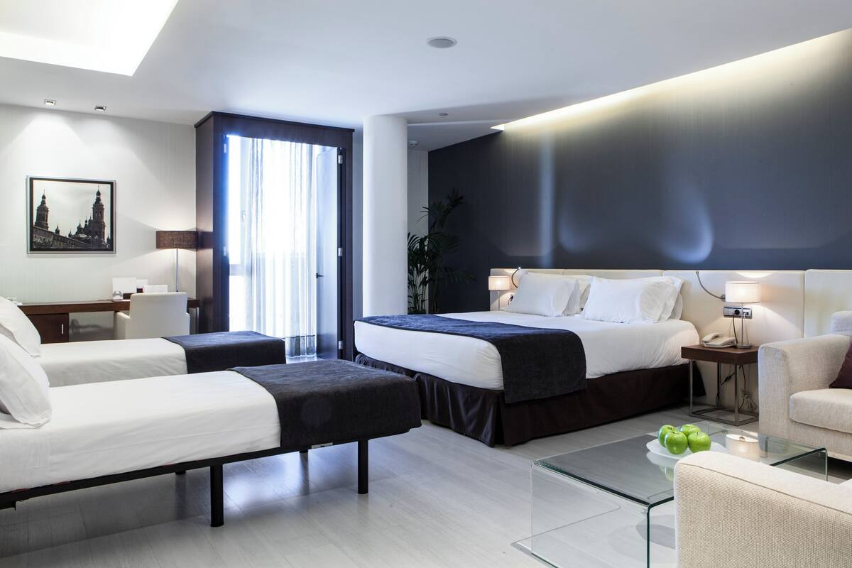 A modern hotel room with a large bed, a sofa, a desk, and a glass coffee table.