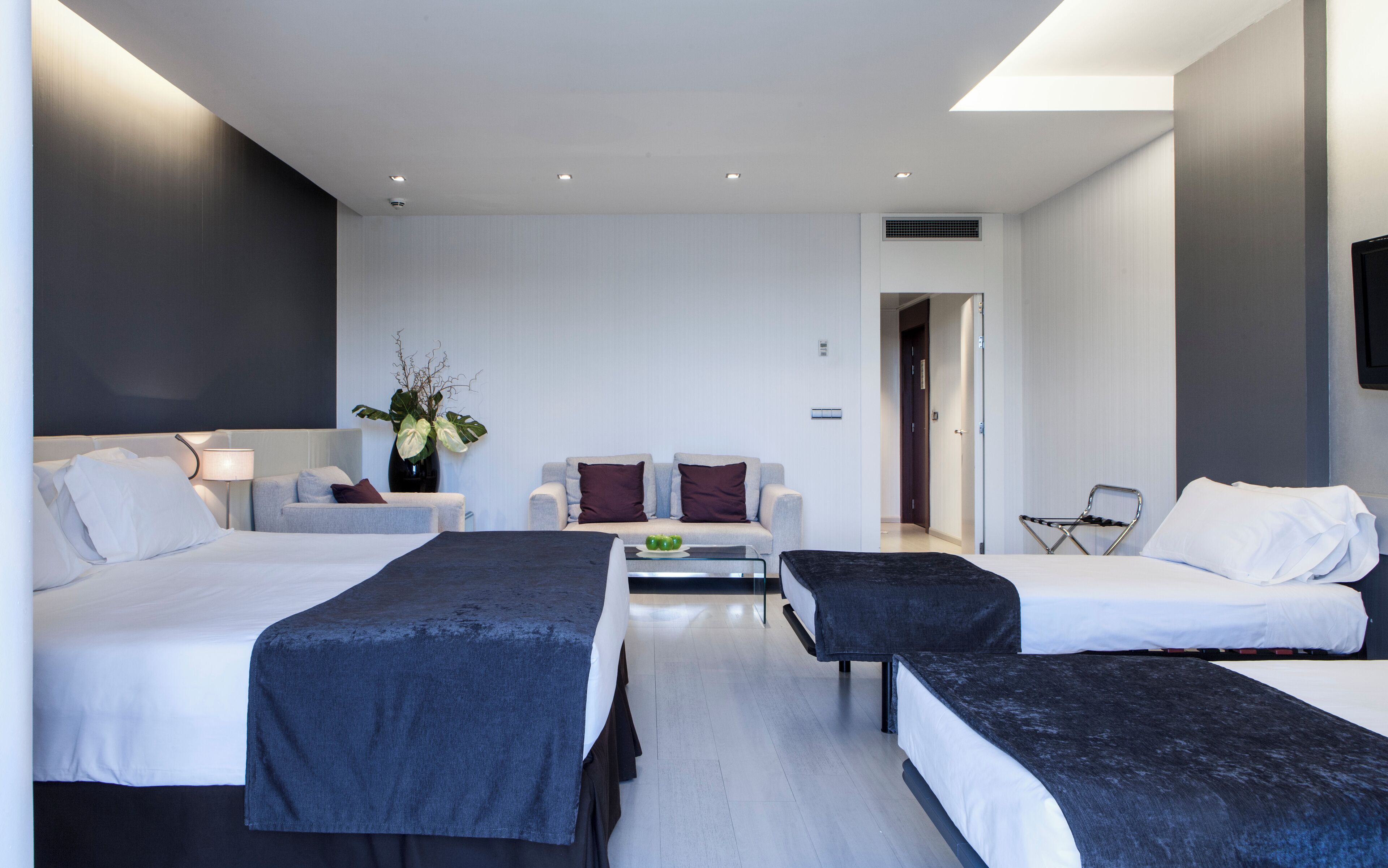 A modern hotel room with two beds, a sofa, a coffee table, and a TV.