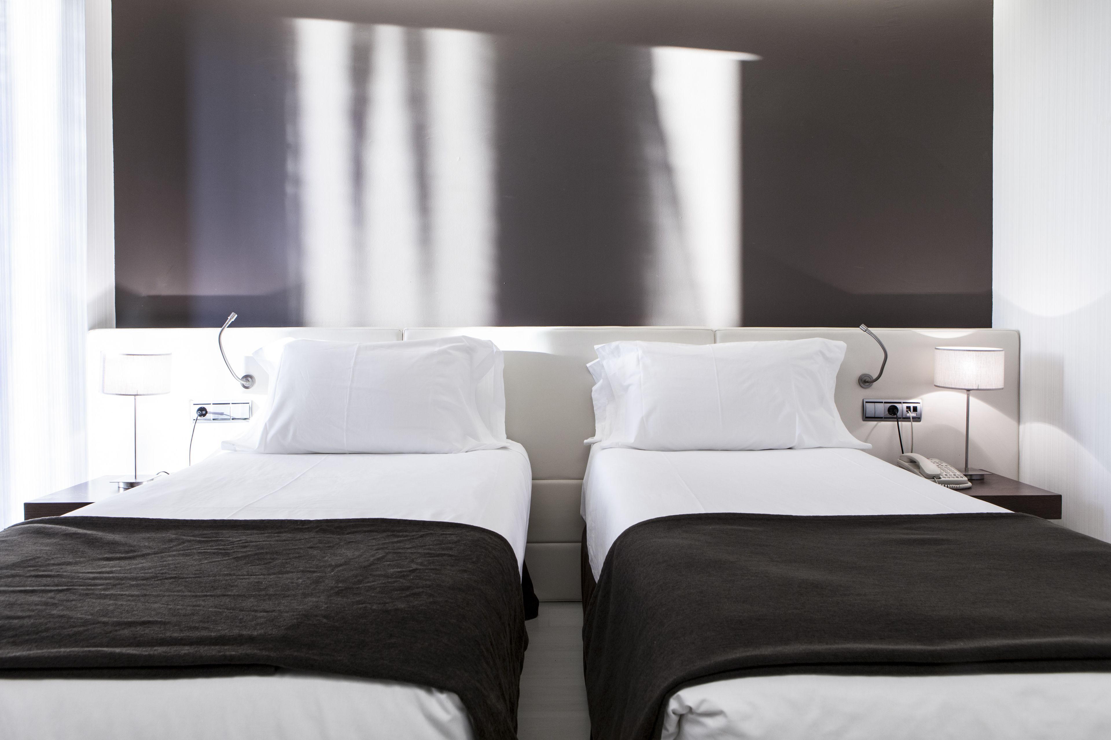 Habitación de hotel con dos camas, cada una con edredón blanco y cubrecama oscuro. Hay mesitas de noche con lámparas y teléfono.