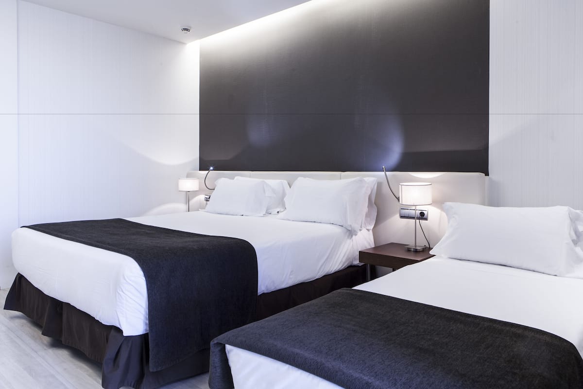 A modern hotel room with two beds, a large dark wall, and a small bedside table with a lamp.