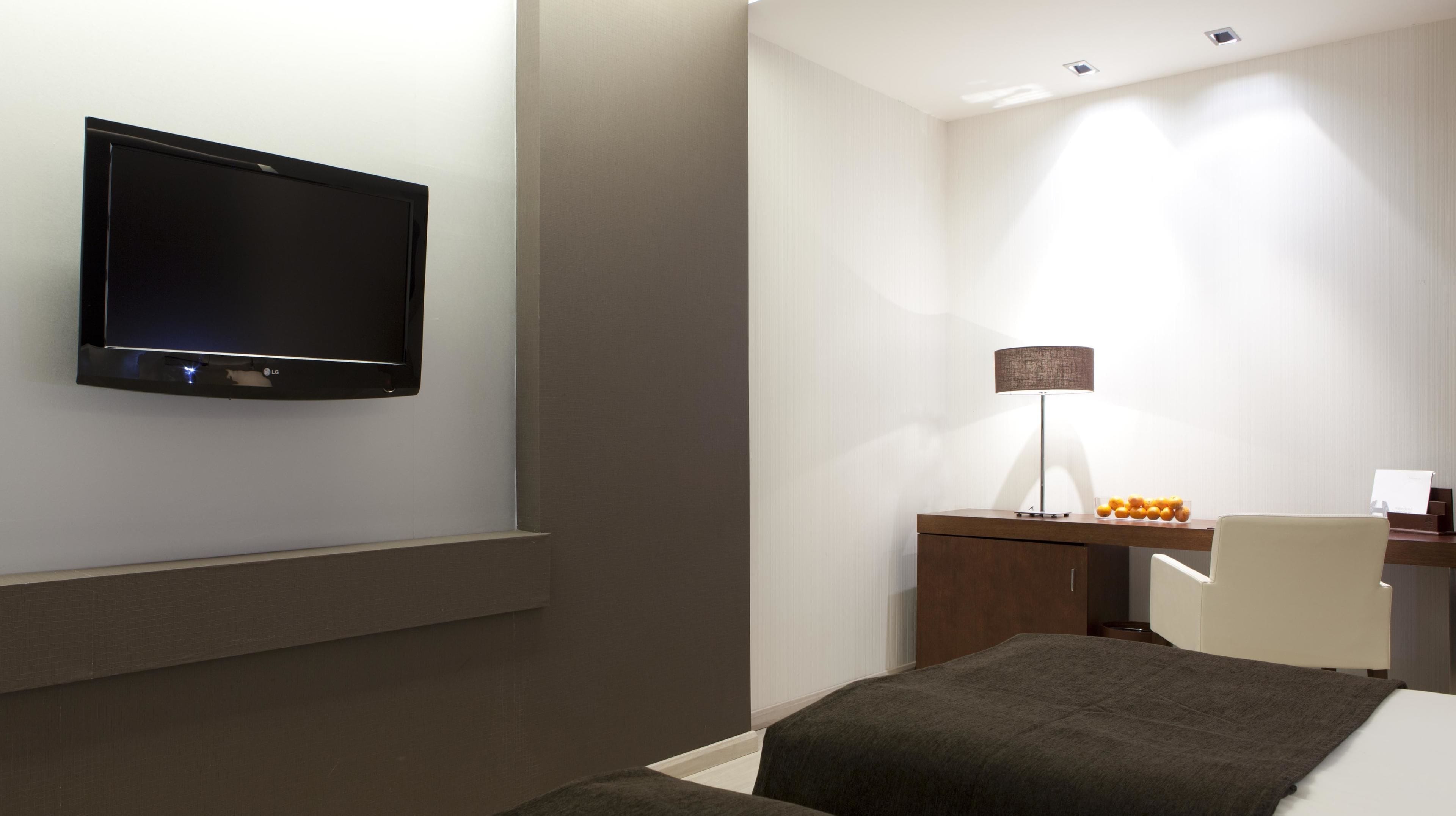 Una habitación de hotel moderna con una televisión de pantalla plana montada en la pared, un escritorio con una lámpara, una silla y una cama con un edredón oscuro.