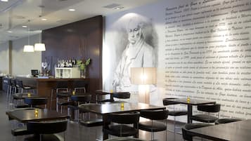 Un restaurante moderno con un gran mural de una figura histórica, mesas con sillas y una zona de bar con botellas y copas.