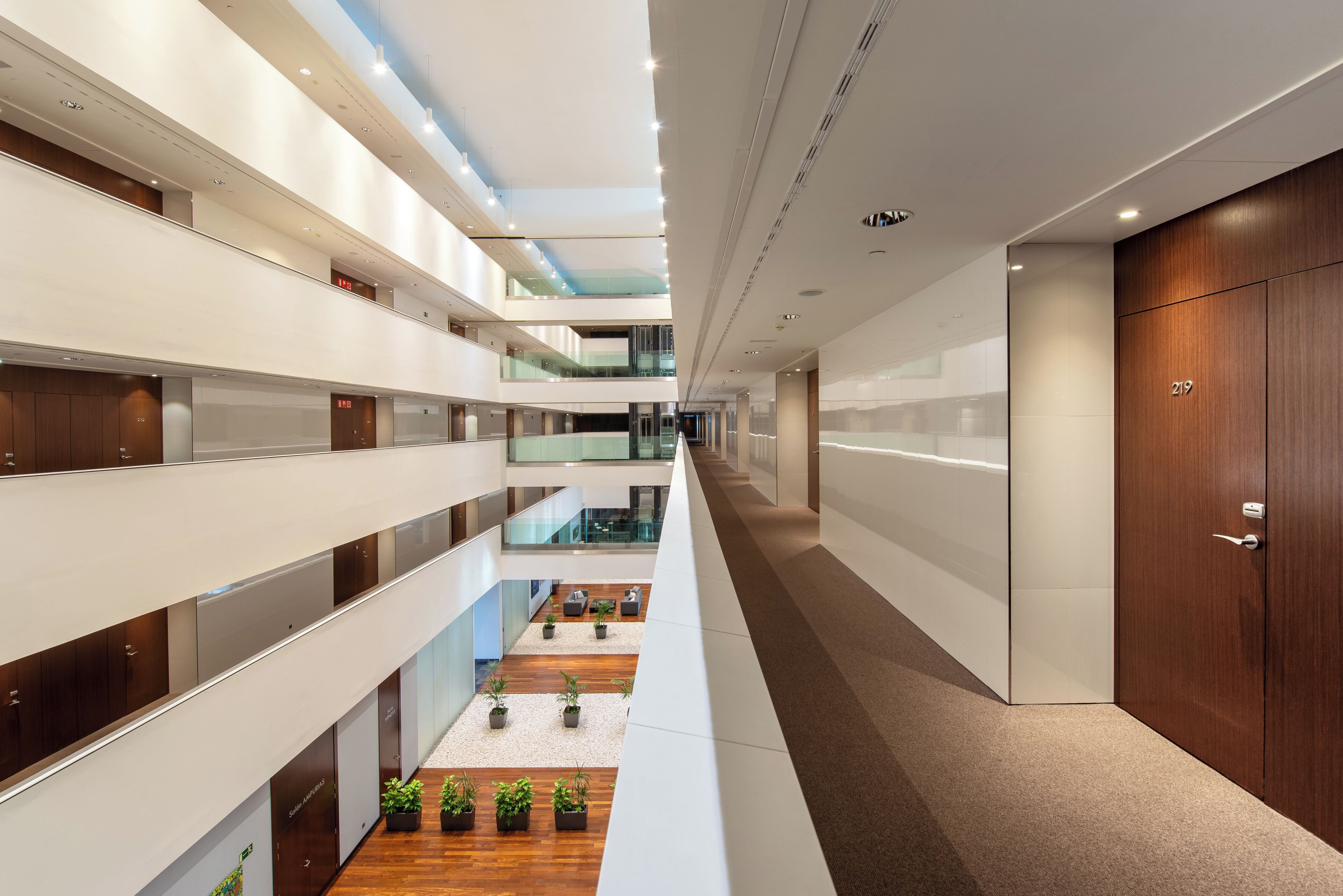 A modern hotel interior with multiple floors, glass railings, and a central atrium with plants.