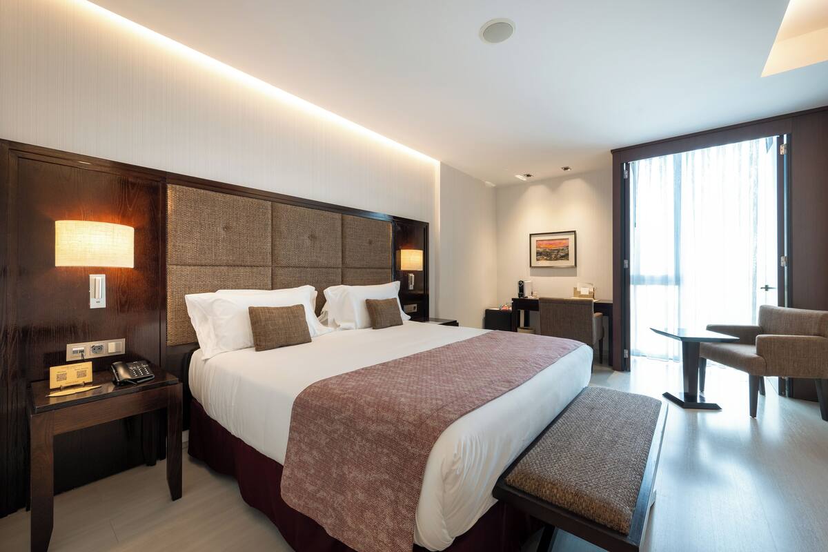 A hotel room with a large bed, bedside lamps, a desk with a telephone, a chair, and a window with curtains.