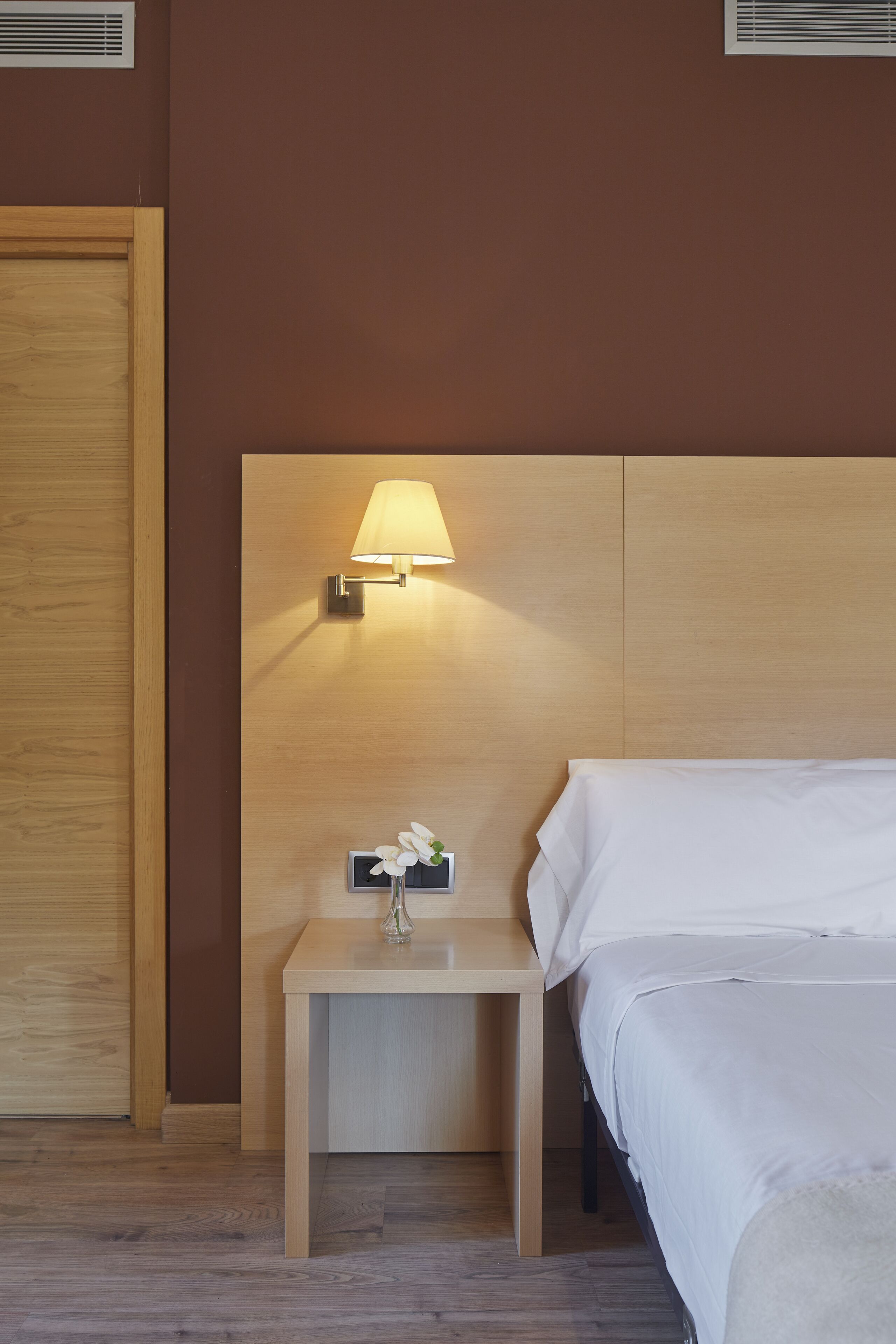 A hotel room with a wooden headboard, a bedside table with a lamp, a small vase with a flower, and a bed with white bedding.