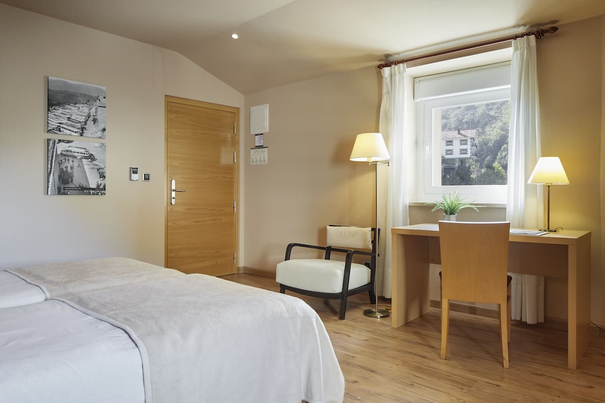 A hotel room with a bed, a desk, a chair, a window with curtains, and two wall-mounted lamps.