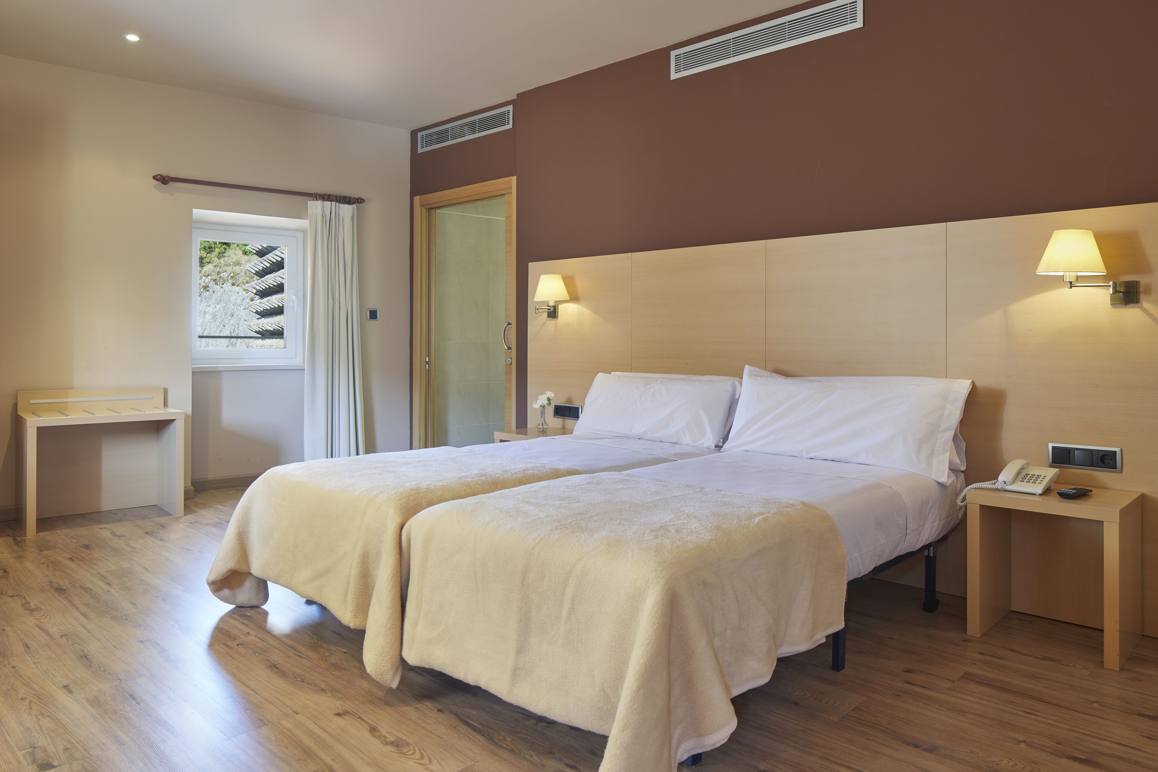 Habitación de hotel con dos camas, piso de madera, ventana con cortinas, mesita de noche con lámpara y teléfono.