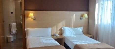 Habitación de hotel con dos camas, suelo de madera, un ventanal grande con cortinas y un baño a la vista.