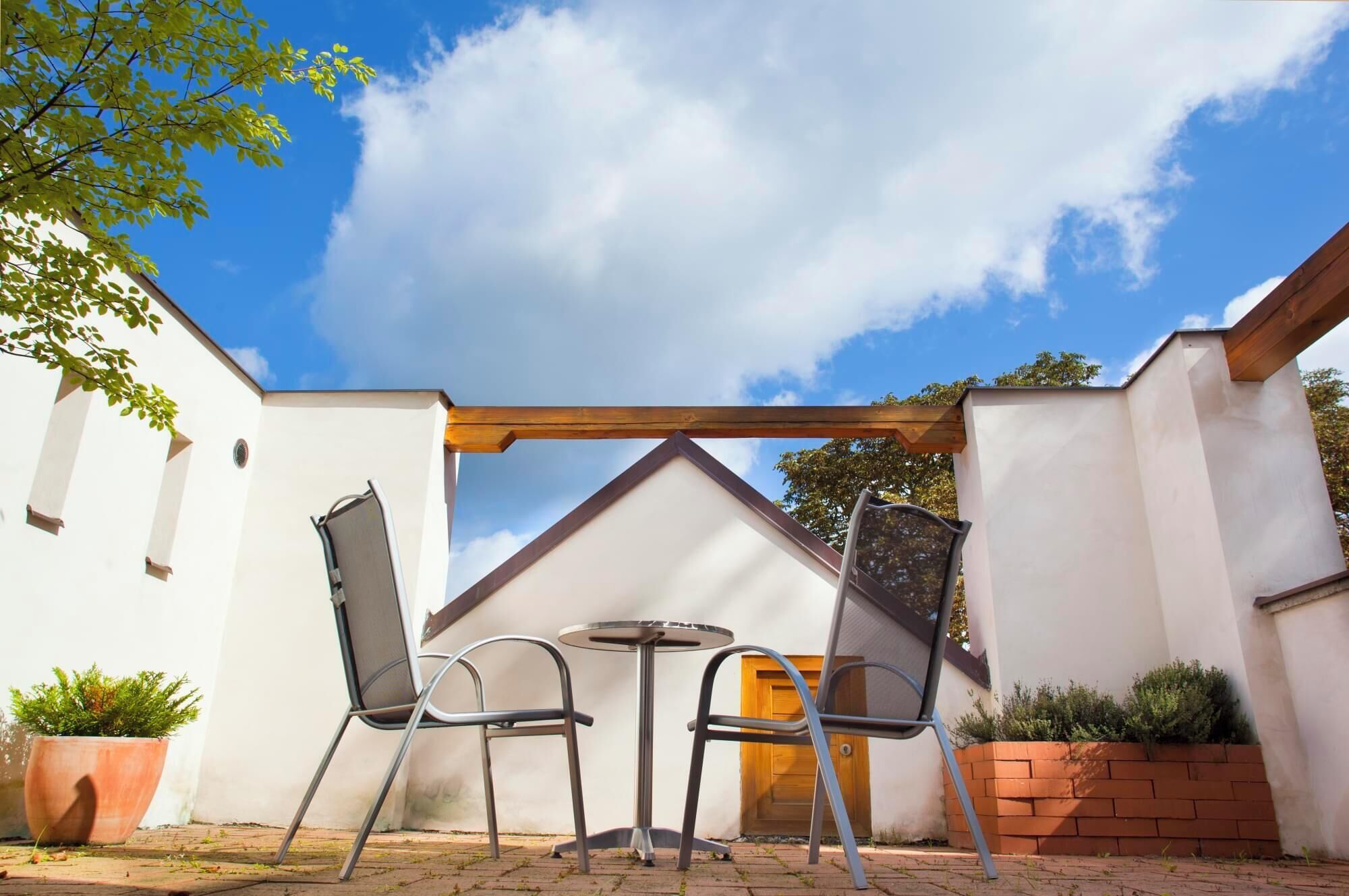 An outdoor patio with two metal chairs and a small round table, a wooden pergola, and a white wall with a window.