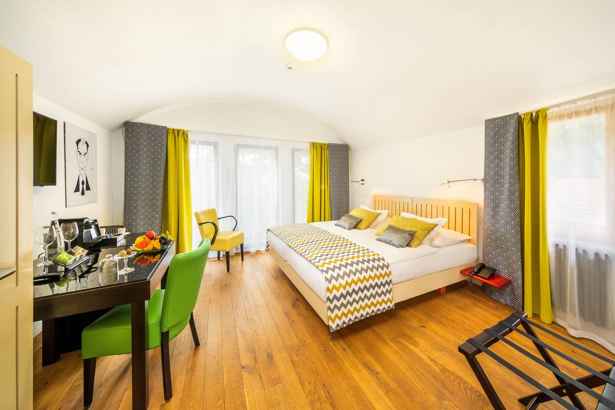 Habitación de hotel con cama, mesa de comedor con sillas, pequeña cocina y ventana con cortinas.
