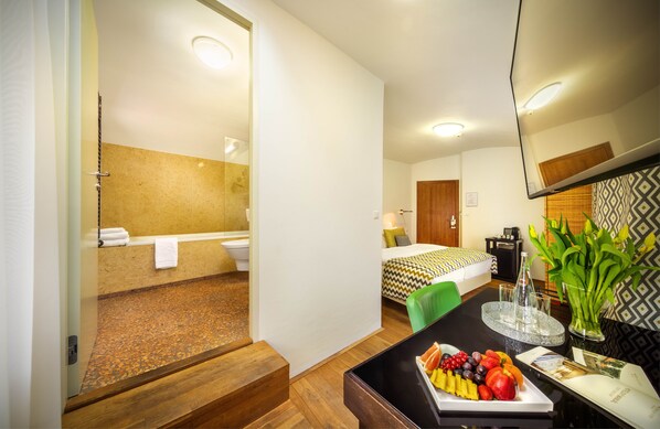 A hotel room with a wooden floor, a bed with a patterned bedspread, a bathroom with a beige wall, and a fruit bowl on the table.