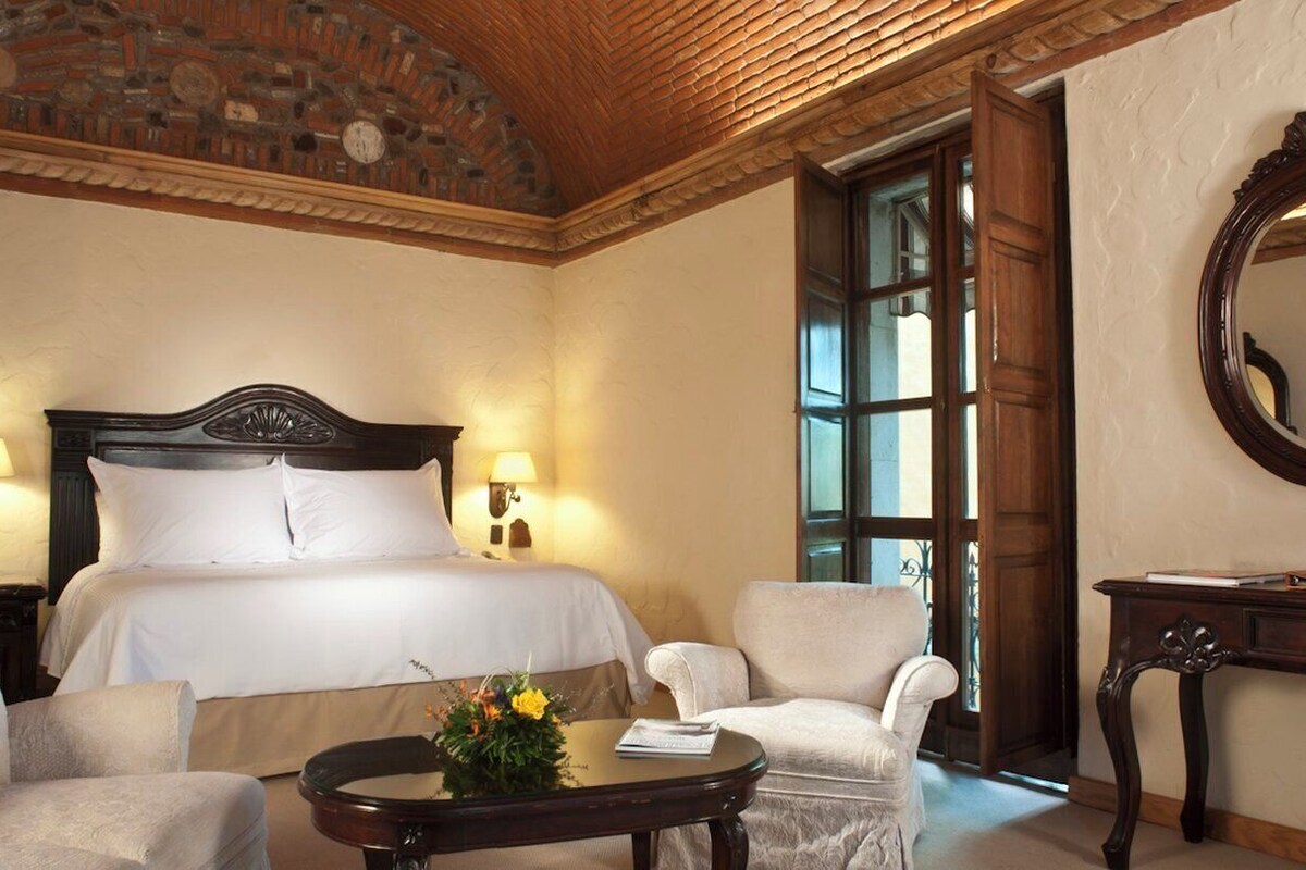 Habitación de hotel con una cama grande, dos sillones, una mesita de noche con un florero y un espejo en la pared.