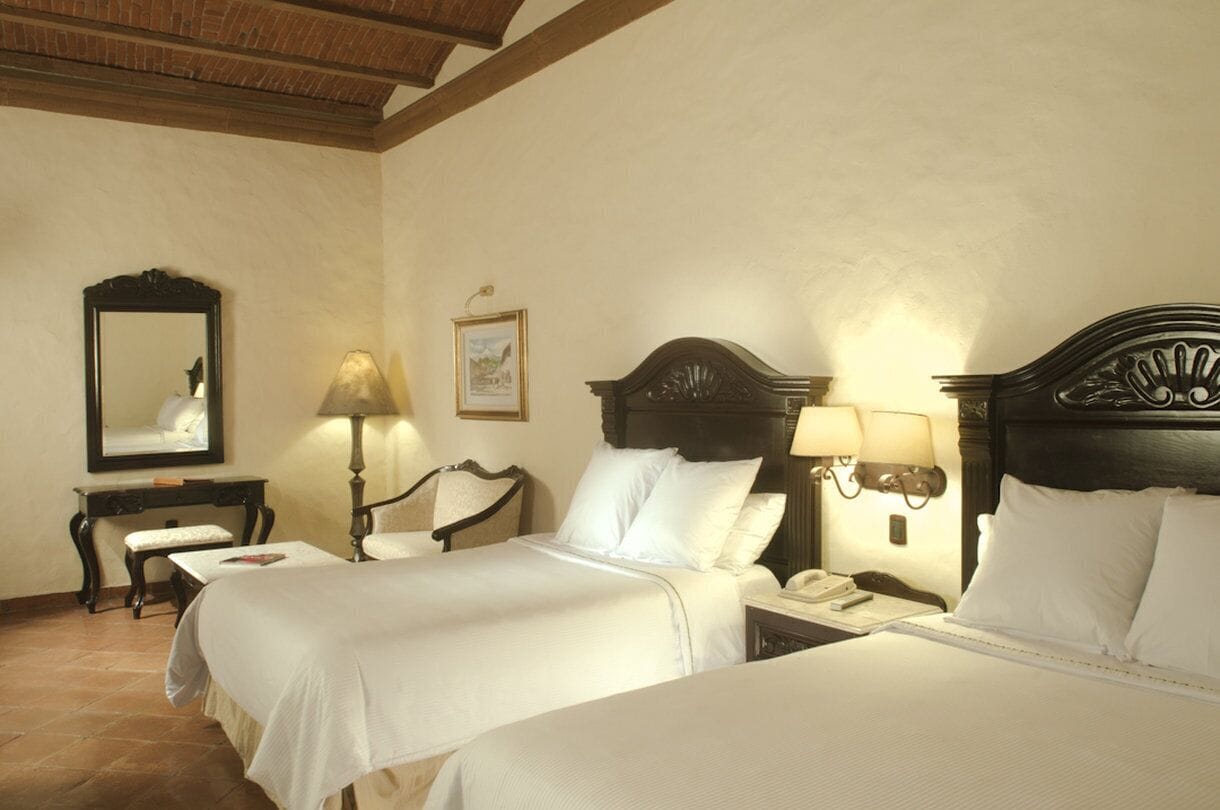 A hotel room with two beds, a desk, a chair, a lamp, and a mirror.