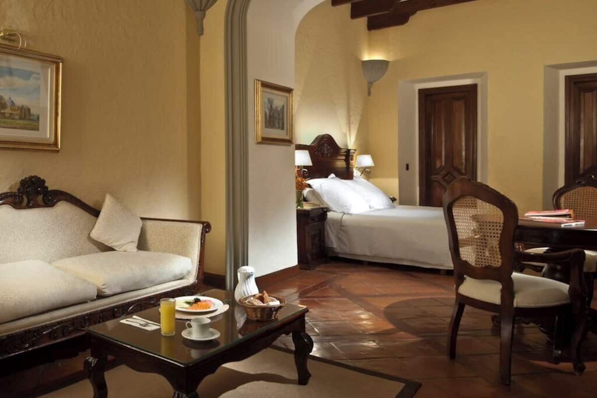 Ein Hotelzimmer mit einer Couch, einem Bett, einem Esstisch mit Stühlen und einem kleinen Tisch mit Tasse und Korb.