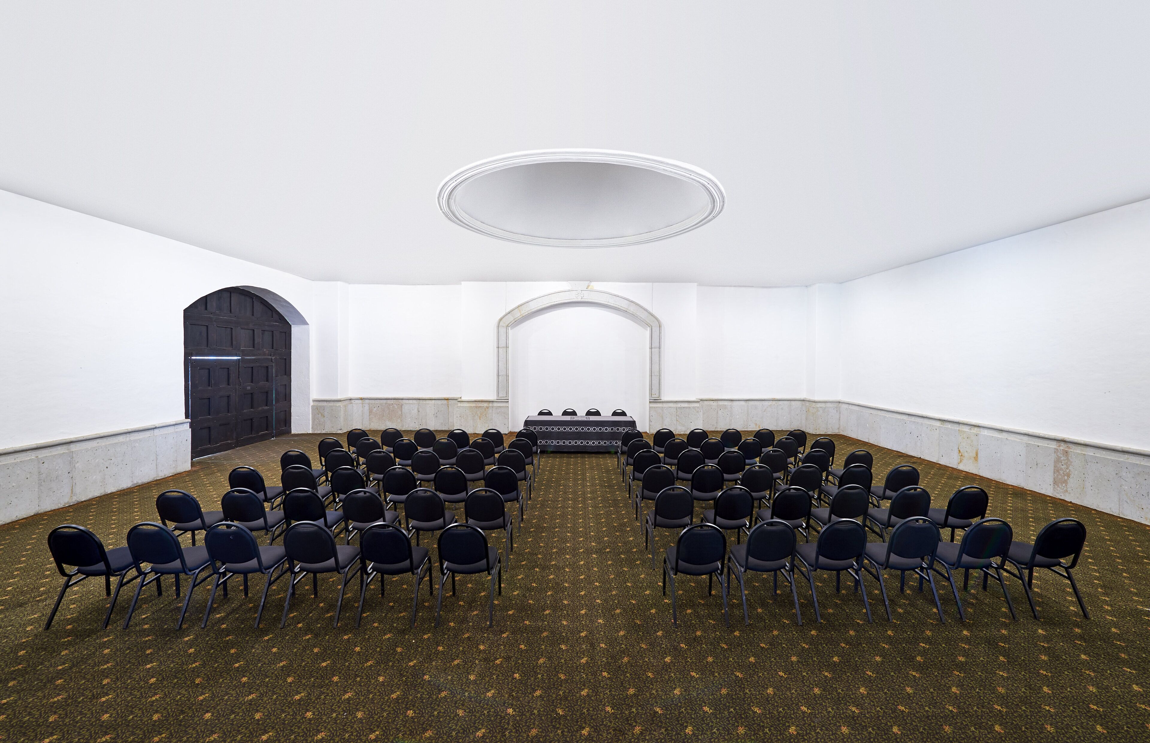 A conference room with rows of chairs facing a stage, white walls, and a carpeted floor.