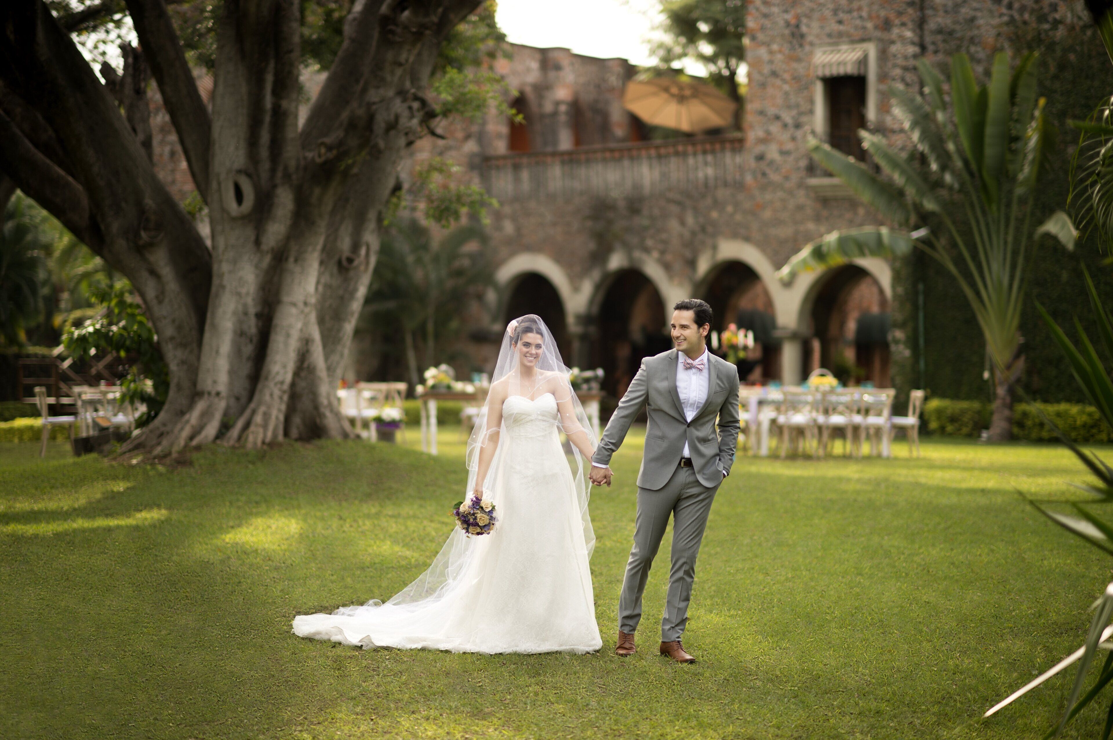 Una pareja tomados de la mano en una zona cubierta de hierba, con un gran árbol y un edificio con arcos al fondo.