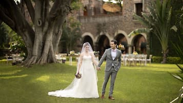 Una pareja tomados de la mano en una zona cubierta de hierba, con un gran árbol y un edificio con arcos al fondo.